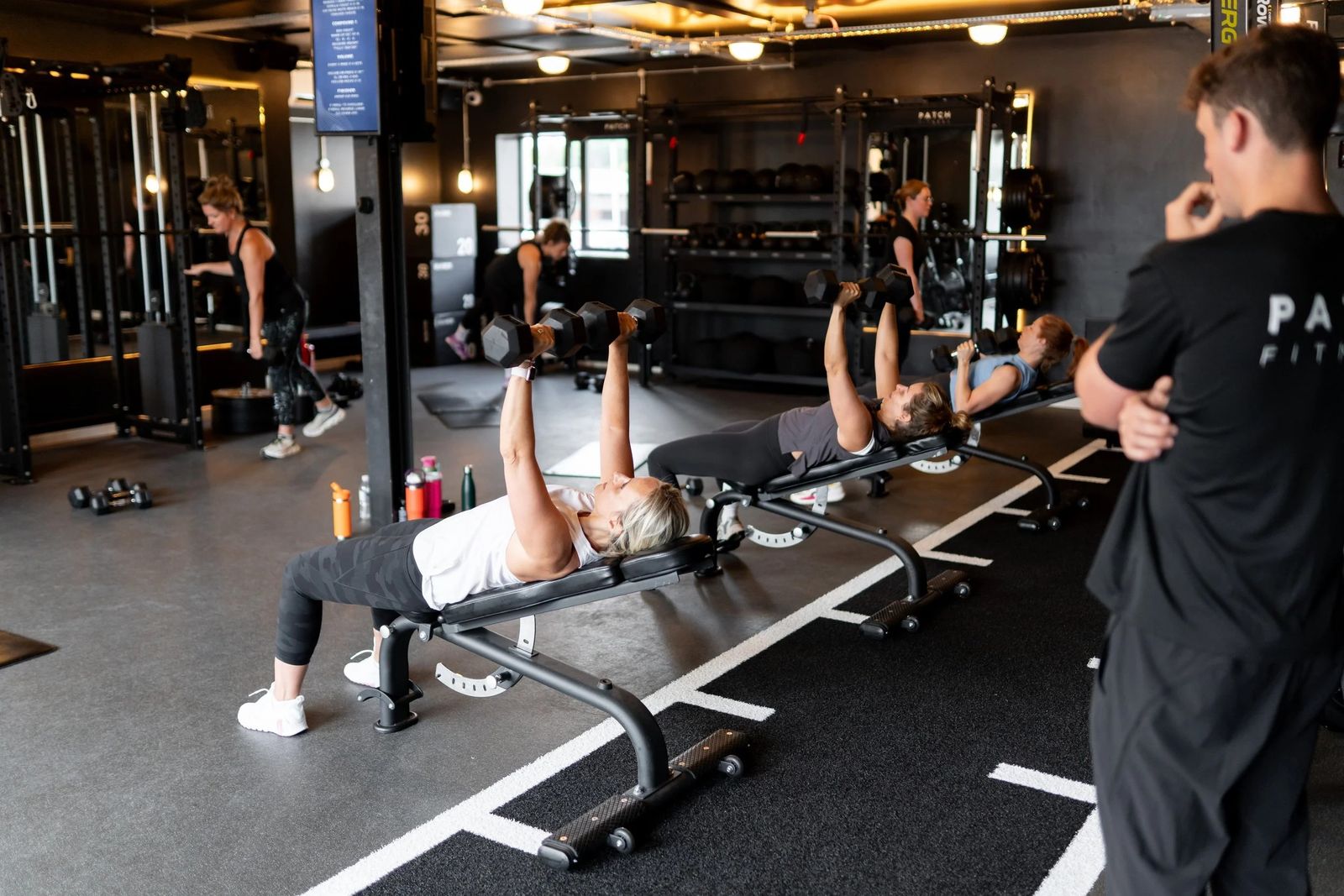 Three members pressing on benches in a row, a coach watching